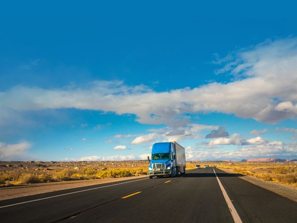 Blue semi-truck driving on a highway under a bright sky.