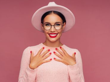 Smiling woman with glasses and pink hat showing red nails.