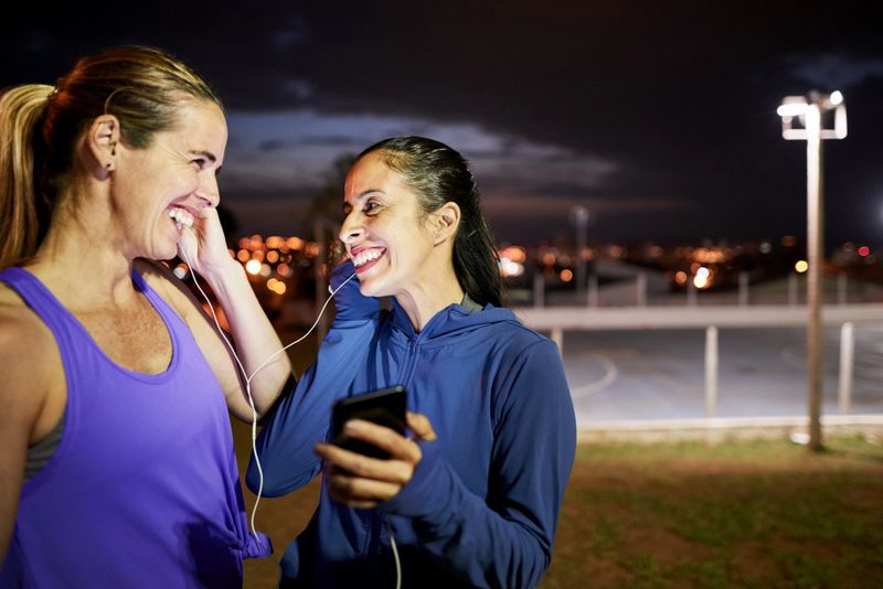 Two friends smiling and looking at each other while listening to the music on smart phone after night run outdoors