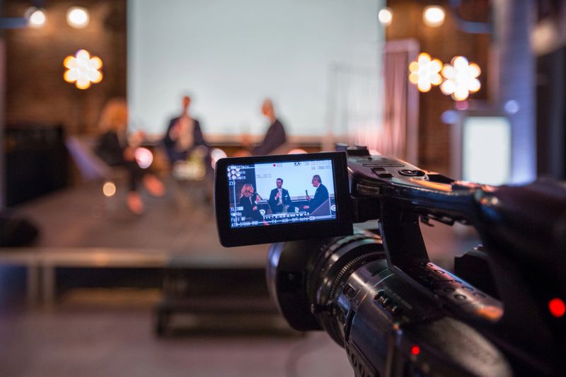 Businesswoman and businessmen discussing during online seminar, sitting on armchairs on the stage. Focus on video camera.