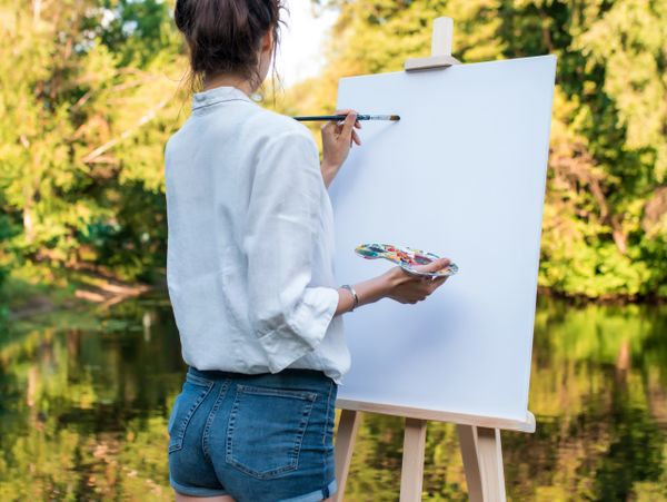 Woman painting on a blank canvas outdoors near a pond.