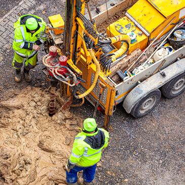 Three construction workers operate a drilling machine on a muddy site.
