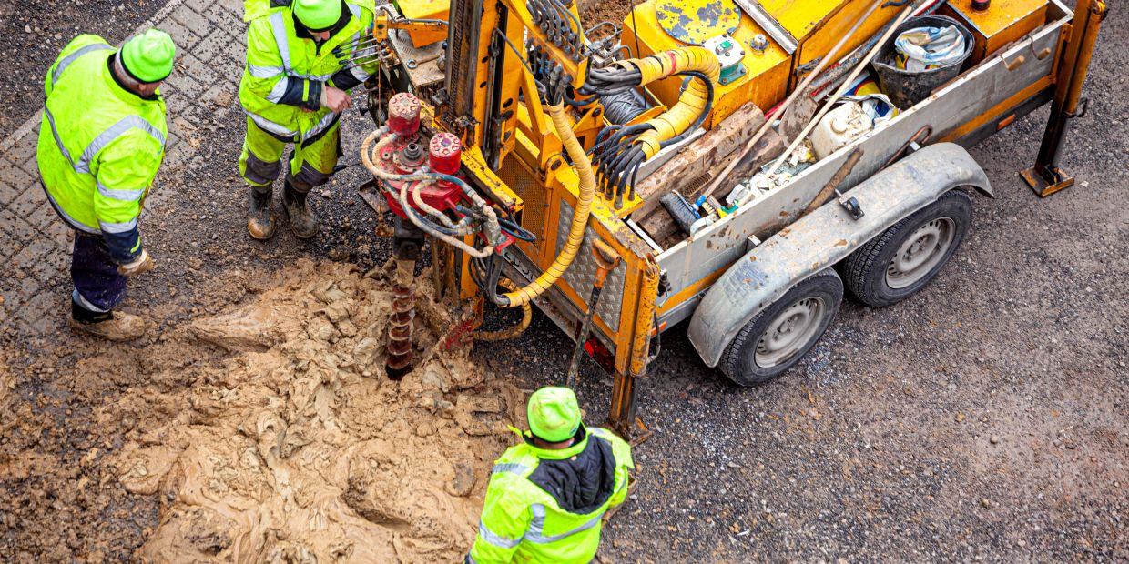 Workers in high-visibility gear operate drilling machinery on muddy ground.