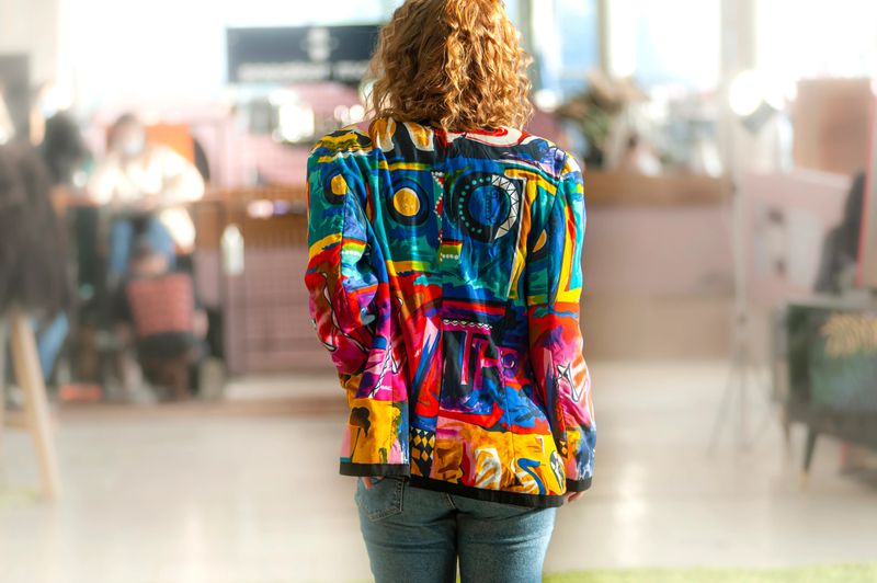 redhead beautiful girl with curly hair and good figure in multicolored jacket with abstract print standing back to camera on unclear background