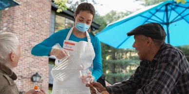 A masked worker serves water to elderly customers outdoors.