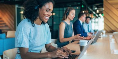 A smiling woman with a headset working on a laptop in a modern office.