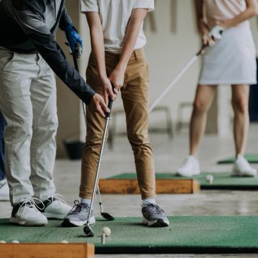 Golf instructor guiding a student on putting technique at an indoor golf practice area.