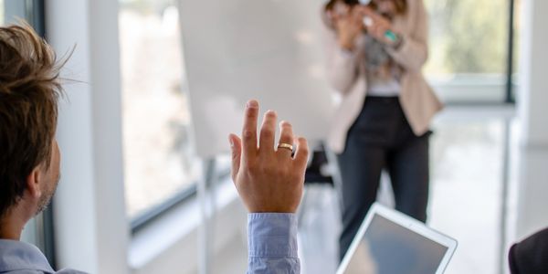 Person raising hand during a meeting with a blurred speaker in the background.