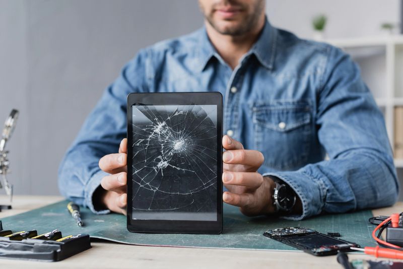 Cropped view of repairman showing smashed digital tablet, while sitting at workplace
