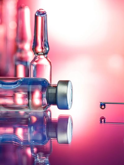 Close-up of vaccine vials and a syringe with a droplet against a pink background.
