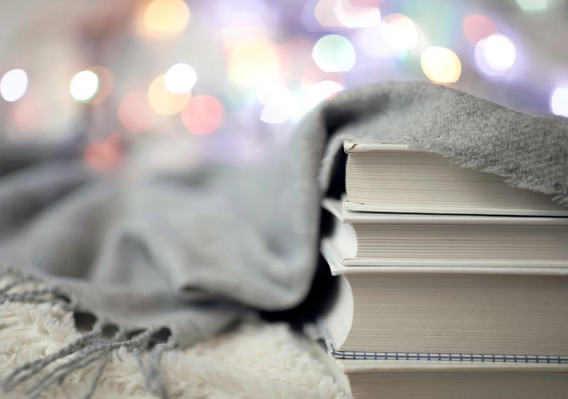 A woolen warm knitted blanket, colorful lights for home decor and a pile of books on the bed. Time to read at home. Cozy and comfortable atmosphere
