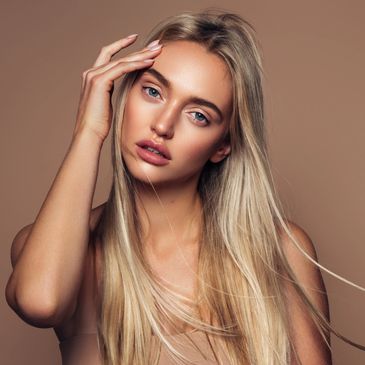 Blonde woman with flowing hair touching her face against a neutral background.