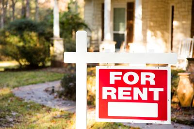 Red and white 'For Rent' sign in front of a house.