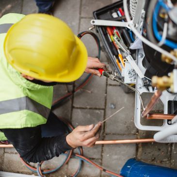 Technician in a yellow hard hat repairing HVAC equipment outdoors.