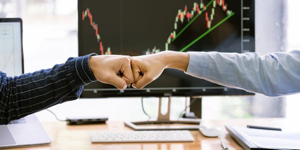 Two people bump fists in an office with stock charts on screens.