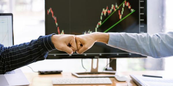 Two people bump fists in an office with stock charts on screens.