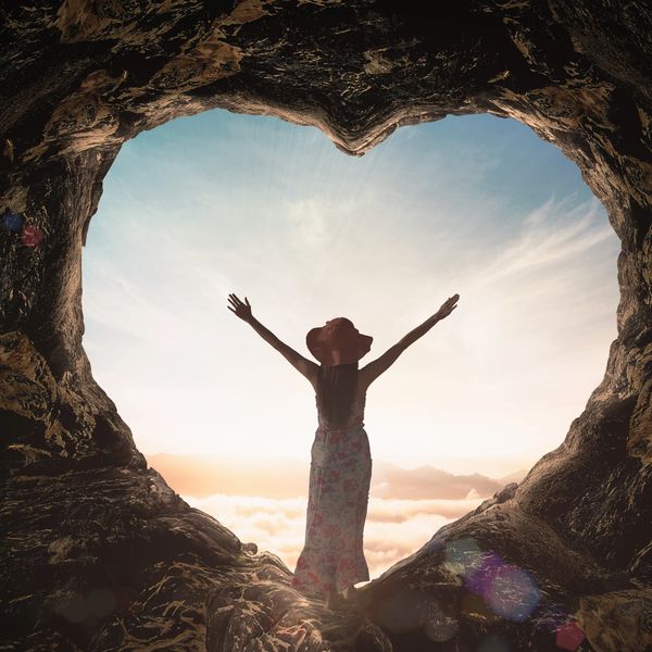 Person standing with arms raised in a heart-shaped cave opening.