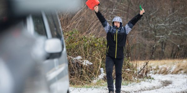 A person signals for help near a snowy roadside with arms raised, holding red and green objects.