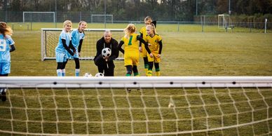 Children in yellow and blue soccer uniforms play on a field with a coach holding a soccer ball.