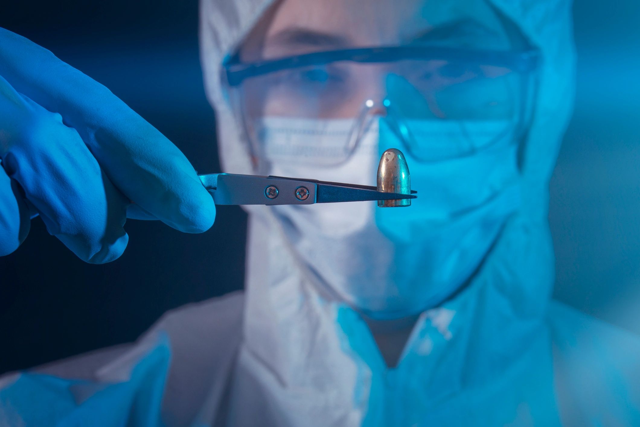 Scientist in protective gear holding a bullet with tweezers.