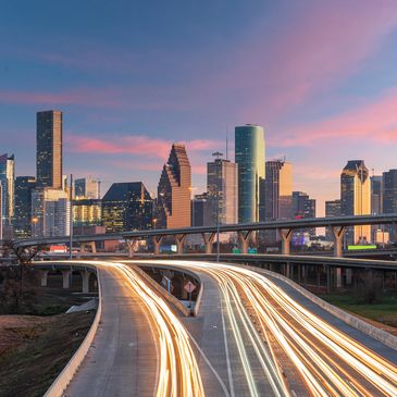 Sunset over a bustling highway leading to the Houston skyline with illuminated skyscrapers.