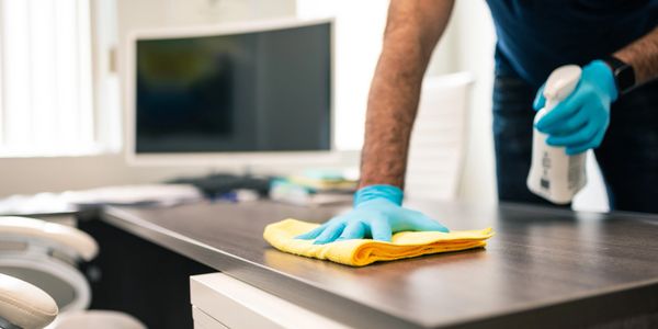 Person cleaning an office desk with a yellow cloth and spray bottle.