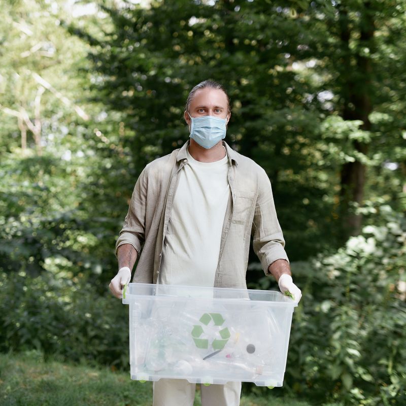 Environmental conservation and ecology. Young caucasian man eco activist wearing protective face mask holding recycle bin and looking at camera while cleaning in the forest during covid 19 outbreak