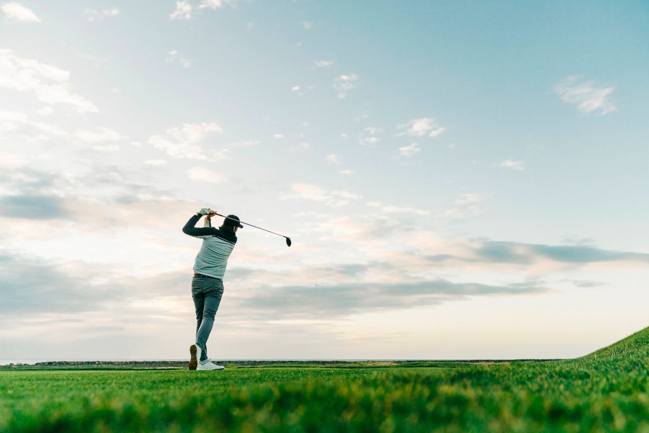 A golfer swings on a lush green course under a cloudy sky.