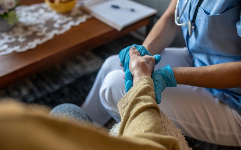 Young nurse in scrub, sitting and holding hand of senior woman. They are in living room, during home care visit. Young nurse is happy to give help to oldest ones.