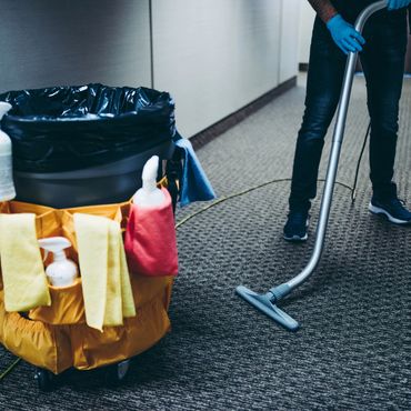 Custodian vacuuming carpet with cleaning cart nearby.