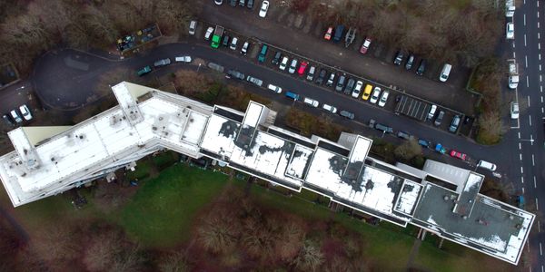 Aerial view of a long white-roofed building with parked cars and surrounding trees.