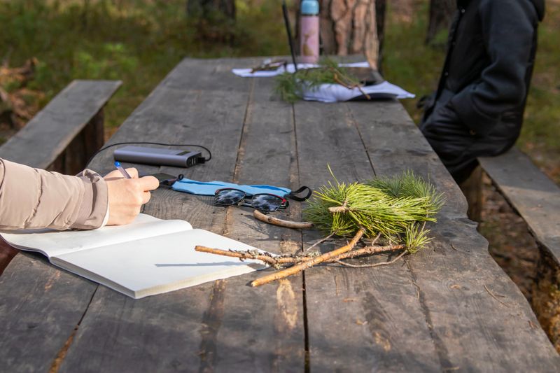 A woman in nature, in a forest, at a wooden table, listens to audio recordings in headphones and writes information in a notebook.