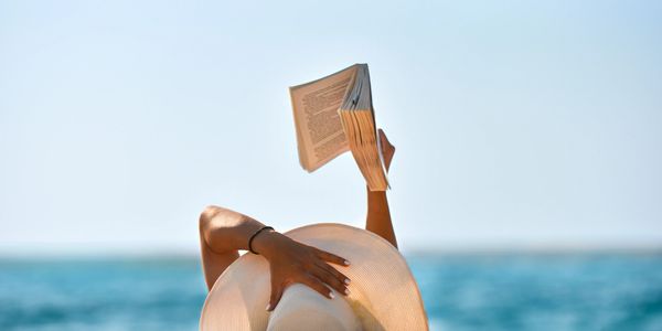 Person lying by the sea, reading a book with a sunhat.