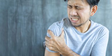 Man in gray shirt clutching his shoulder in pain with eyes closed.