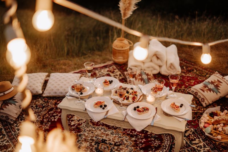 Served Picnic Table with Tasty Desserts and Drinks Outdoor Evening Dinner Party. Fruits and Berries, Cheese and Drink on Vintage Desk. Glowing Lamps and Decorations. Towels and Pillows on Carpet