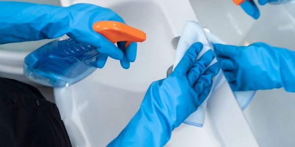 Hands in blue gloves cleaning a white sink with spray and cloth.