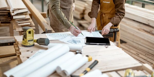Two workers discuss architectural plans in a woodworking shop.