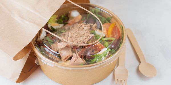 A fresh salad with tuna in a clear plastic container beside wooden utensils.