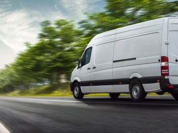 A white delivery van driving on a tree-lined road.