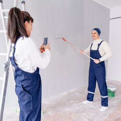 Couple painting a wall, woman taking photo of man with paint roller.