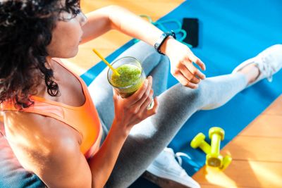 Woman on exercise mat drinking green smoothie with dumbbells nearby.