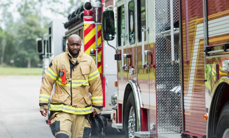 An African-American firefighter wearing a fire protection suit and carrying a helmet, walking beside two fire engines. He is looking at the camera with a serious expression. He is a young man in his 20s.
