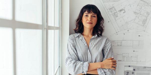 Confident woman stands with arms crossed by architectural blueprints near a bright window.