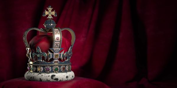 An ornate royal crown with jewels on a red velvet cushion.