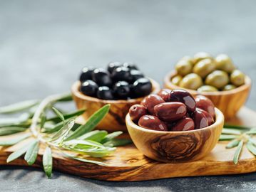 Three wooden bowls filled with black, green, and purple olives on a wooden board.