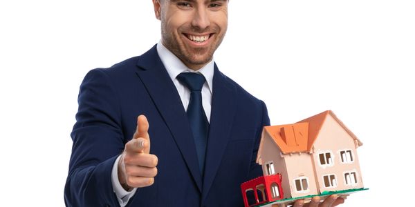 Smiling man in suit holding a house model and pointing forward.