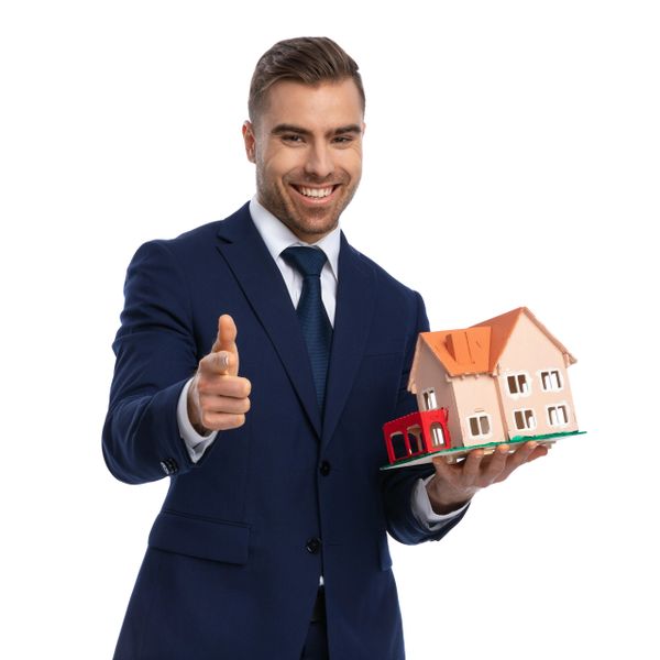 Smiling man in suit holding a house model, pointing at camera.