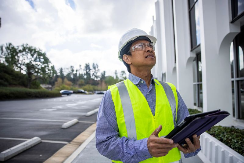 A mature Filipino man with a tablet and safety vest, helmet and glasses inspects a building and takes notes.