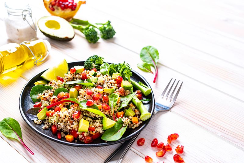 Vegan food backgrounds: healthy fresh multicolored quinoa salad plate shot on white table. The salad includes chopped carrot, avocado, pomegranate, broccoli, red chard leaves and tomato. Ingredients are around the plate. The composition is at the left of an horizontal frame leaving useful copy space for text and/or logo at the right. High resolution 42Mp studio digital capture taken with SONY A7rII and Zeiss Batis 40mm F2.0 CF lens