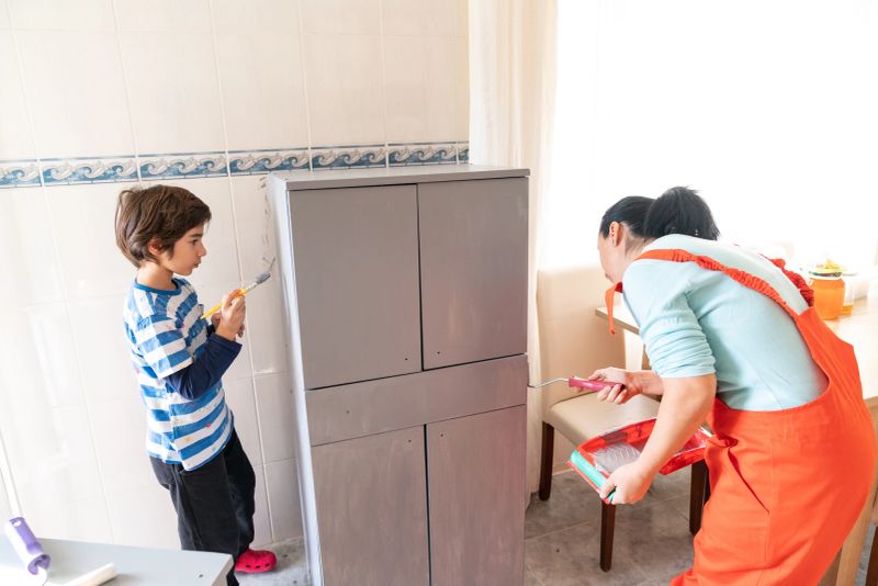 Photo of mother wearing an orange coveralls and son painting kitchen cabinet in the kitchen. They are painting in gray color. Shot indoor with a full frame mirrorless camera.